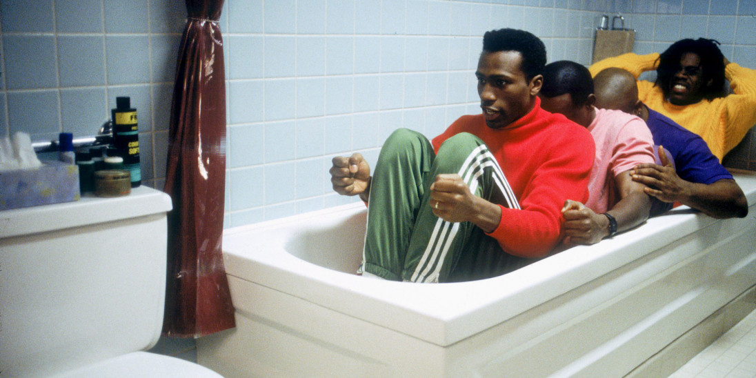 Four men sit fully-clothed in an empty bathtub practicing bobsledding.