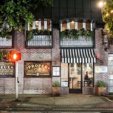 A nighttime photograph of a brick building with glass windows and striped awnings. A crosswalk signal with a red "Stop" symbol is visible to the right of the building's entrance.