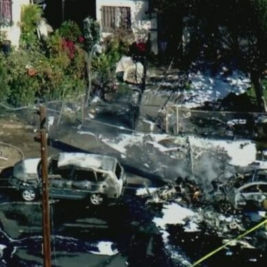 An aerial shot of a neighborhood where a small plane has crashed, leaving two cars burned. Firefighters survey the scene after putting out the flames.